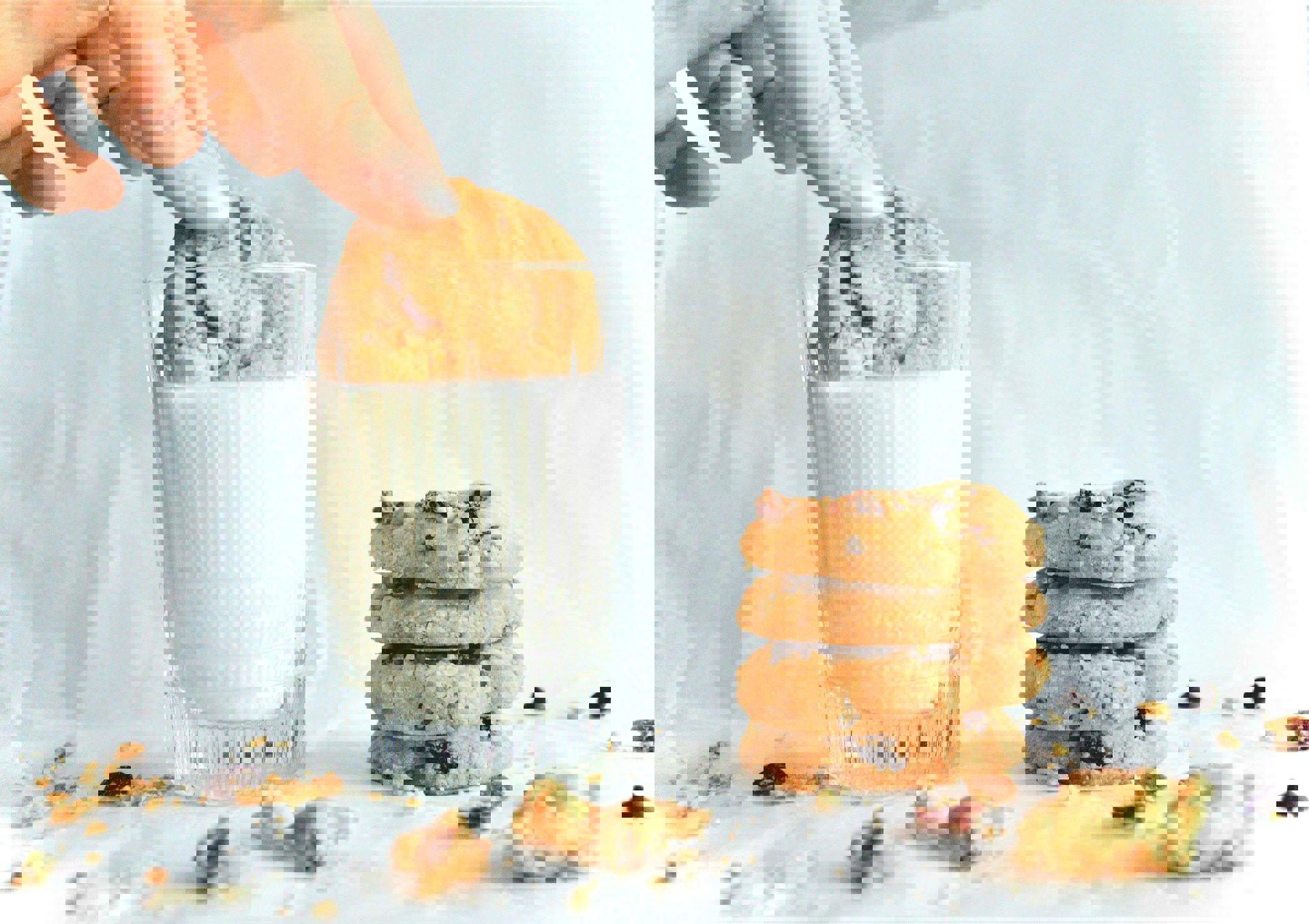 En hånd dypper en småkage i et glas mælk, mens en stak af fire småkager ligger ved siden af på et hvidt underlag med krummer omkring.